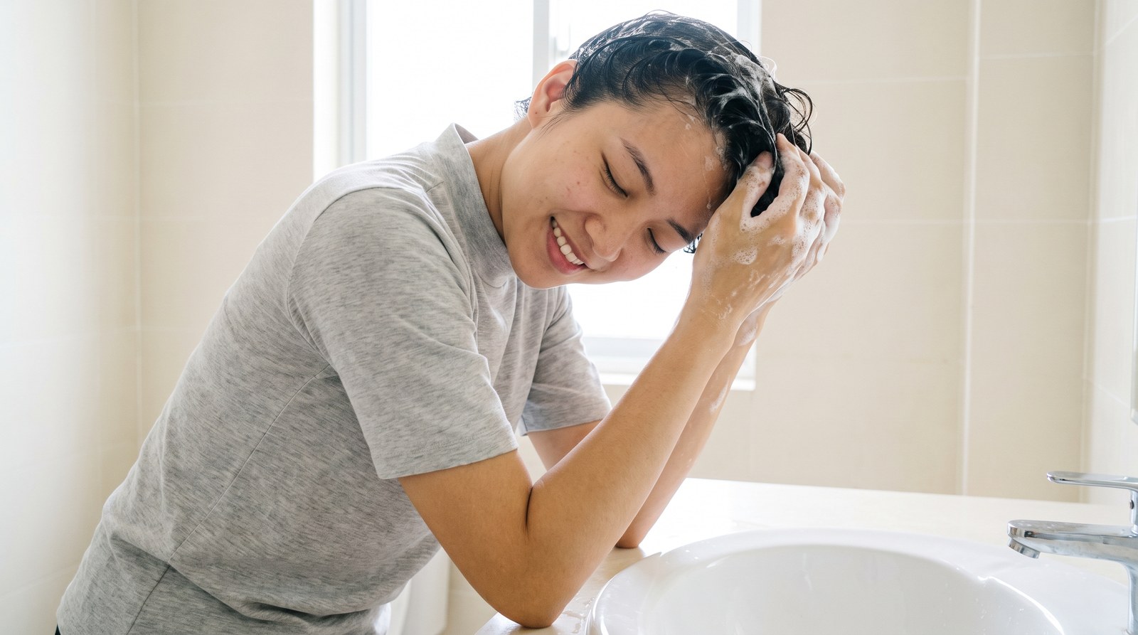 Cô gái Việt trẻ massage da đầu bằng đầu ngón tay trong lúc gội đầu với bọt dầu gội