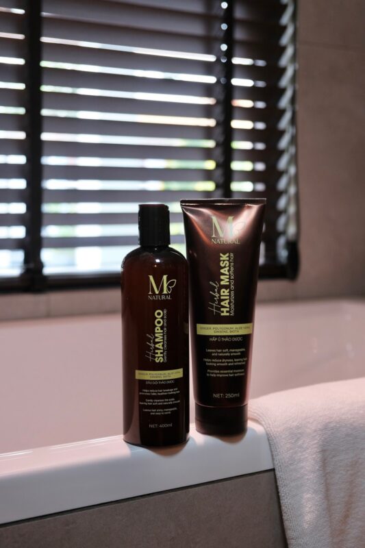 Chai dầu gội và tuýp hấp ủ M Natural bên rèm sáo phòng tắm