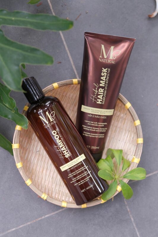 Chai dầu gội và tuýp hấp ủ M Natural trên khay gỗ cùng lá xanh