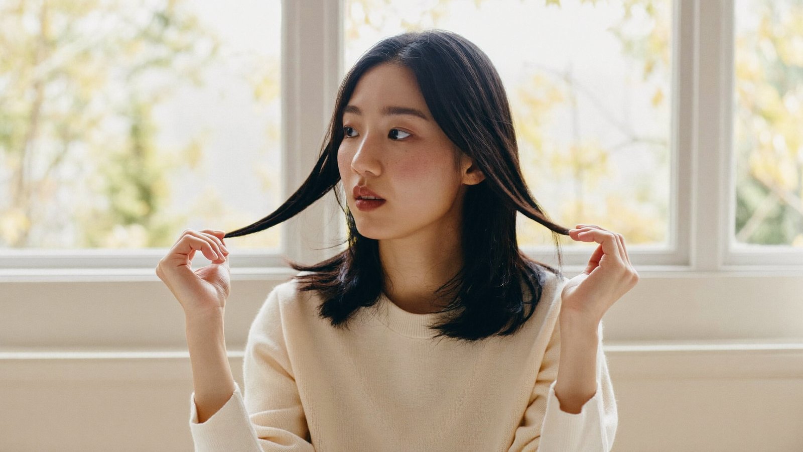 Cô gái trẻ ngồi bên cửa sổ mùa thu, nhẹ nhàng vuốt lọn tóc khi thời tiết chuyển mùa